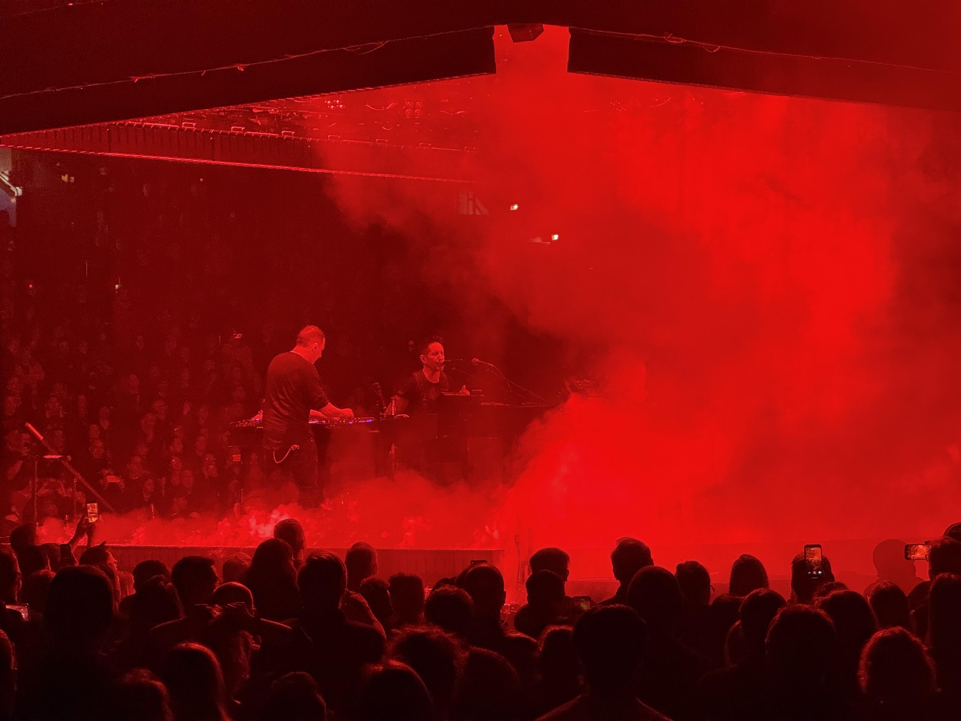 Trent Reznor singing into a microphone on a small stage surrounded by people, shrouded by fog and lit by a red light.