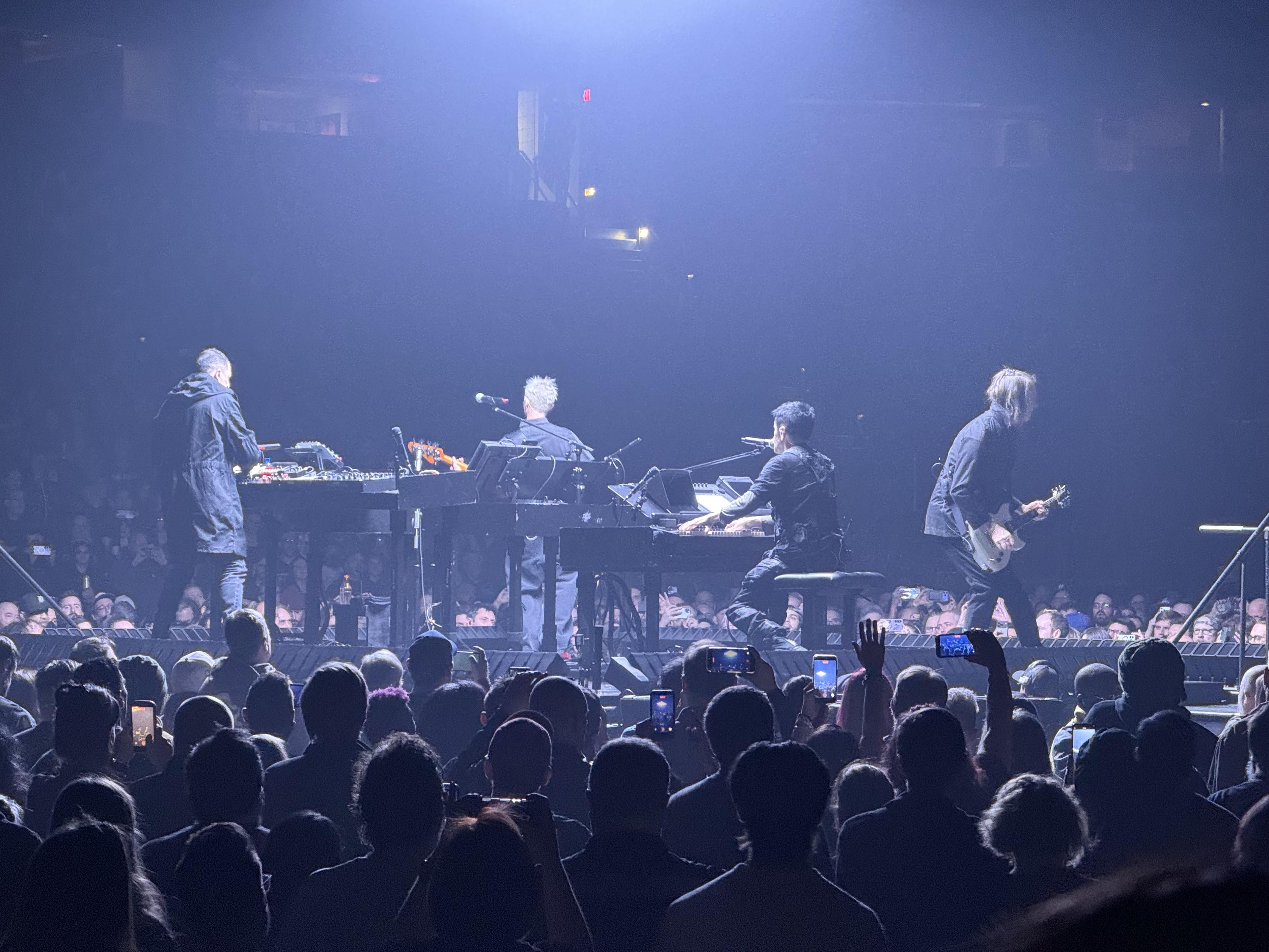 Nine Inch Nails performing on the “B” stage, elevated over a crowd surrounding it on all sides. Four people are playing musical instruments; a synthesizer, bass, guitar, and Trent Reznor is seated at a keyboard piano with his back to the camera.